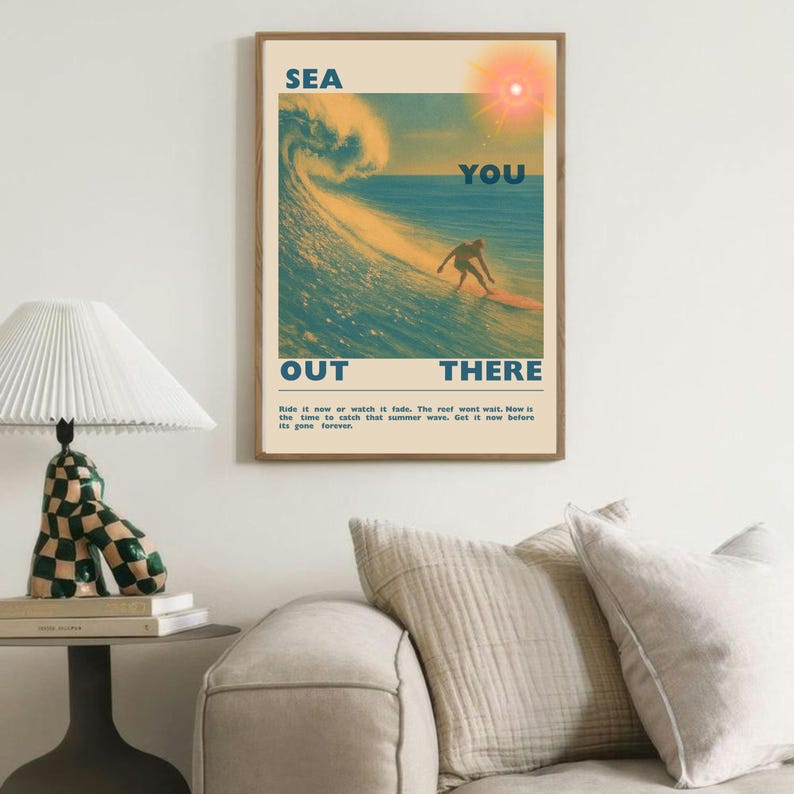 May include: A framed poster with a surfing theme. The artwork features a surfer riding a wave, with the words "SEA YOU OUT THERE" in bold letters. The color palette includes shades of blue, yellow, and orange.