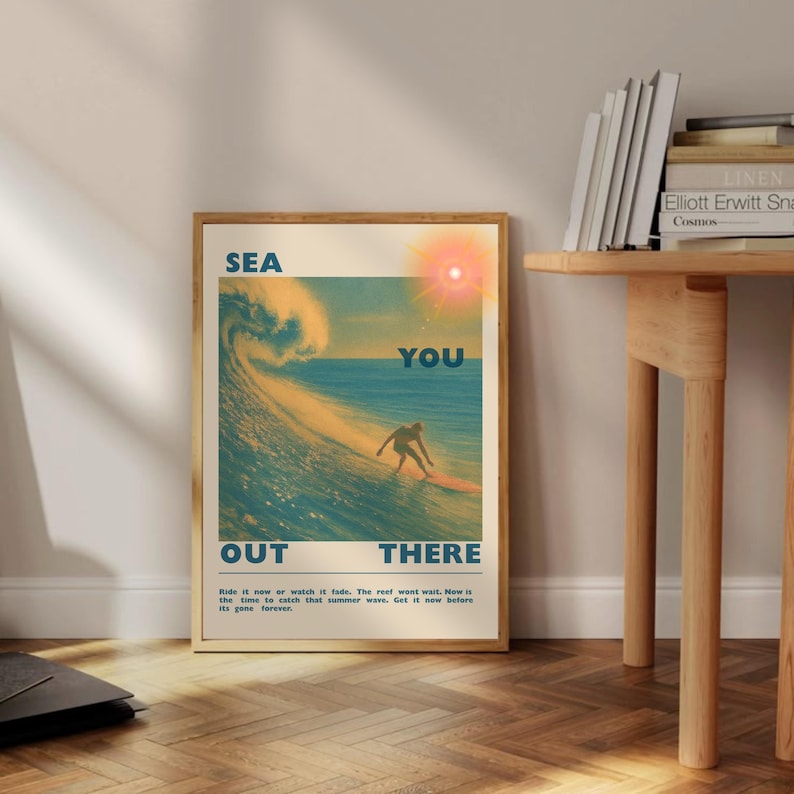 May include: A framed vintage-style poster with a surfing theme. The image features a surfer riding a wave, with the words "SEA YOU OUT THERE" in bold letters. The poster has a warm color palette with a sun and ocean scene.