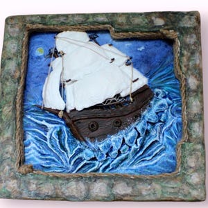 May include: A 3D paper mache artwork depicting a ship sailing through stormy seas. The ship is white with brown sails and a brown hull. The waves are blue and white and are textured to look like water. The artwork is framed in a brown frame with a rope border.