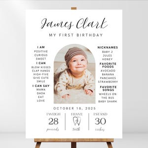 May include: A white poster with black text celebrating a baby's first birthday. The poster includes the baby's name, James Clark, and details about their first year, including their weight, height, and favorite foods and songs. A photo of the baby is featured in the center of the poster.