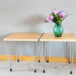 May include: Two light wood end tables with hairpin legs. The tables are next to each other and a vase with pink flowers is on the table on the right.