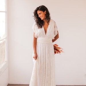 A woman wears a wedding dress with white walls at the back and hydraulic floor. She is wearing a femenine wedding dress made of ivory lace. She also wears a bouquet in the hands, which are in her back. She has long dark boho hair.