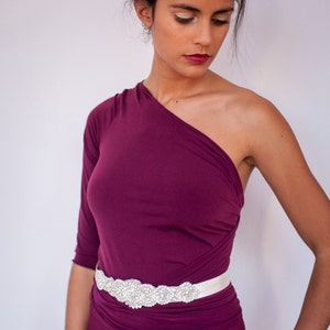 Puede incluir: Un vestido de un solo hombro en color ciruela intenso con un corpi&ntilde;o ajustado y tela fruncida en la cintura. Una faja ancha de sat&eacute;n blanco est&aacute; atada a la cintura, adornada con un panel decorativo central de pedrer&iacute;a y cuentas transparentes.