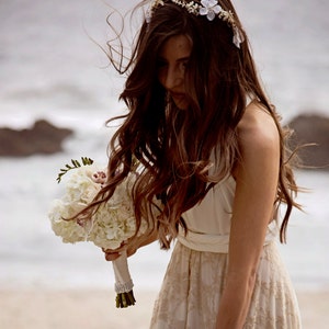 A woman is wearing a golden lace gown beach wedding dress. She is at the beach. The ambiance is dreamy and romantic, and she looks happy. She wears a bouquet in her arms. She wears a flower crown and has dark hair.