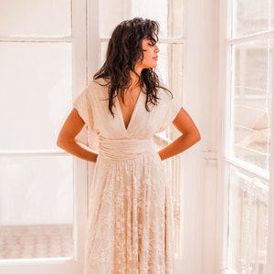 We see a woman next to the window wearing a boho wedding dress. The dress has an ethereal appearence. The woman has dark long hair and is looking to the side. The dress is floor lenght, giving a bohemian and romantic vibe.