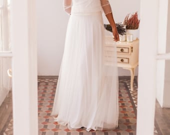 May include: A woman in a white wedding dress with a sheer polka dot top and a flowing skirt. She is smiling and holding a bouquet of flowers.