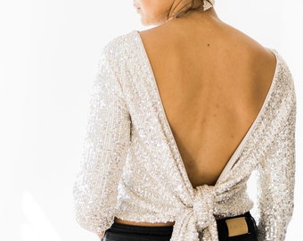 May include: A long-sleeved silver sequin top featuring a deep V-shaped open back with a large fabric tie closure at the waist. The garment is paired with a matching silver sequin headband and black denim jeans with a visible tan leather patch.