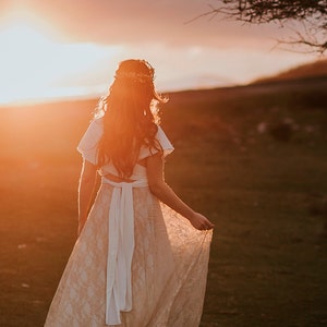 Bride wearing with happiness her boho wedding dress. She is in a landscape that reminds her of Ireland. It's the golden hour and the sun looks incredible.  She is holding the golden lace overskirt of her bridal gown.