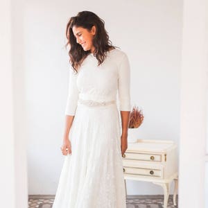 The ivory lace wedding dress looks stunning on the woman. She is a model dressed like a bride. The gown combines a convertible dress with a lace overskirt. She is in front of  drawer