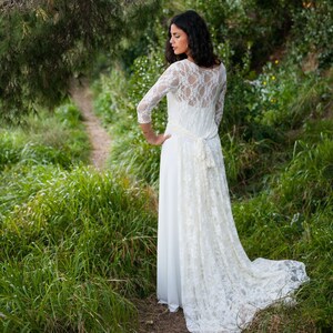 May include: A floor-length white dress featuring three-quarter length lace sleeves, a sheer lace back panel, and a long lace train that attaches at the waist with a tie. The dress has a solid white skirt and a delicate floral lace overlay.