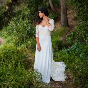 May include: A floor-length white dress featuring three-quarter length lace sleeves, a scoop neckline, and a gathered waist detail. The bodice is constructed from lace, while the skirt is made of a smooth, solid white fabric with a lace train.