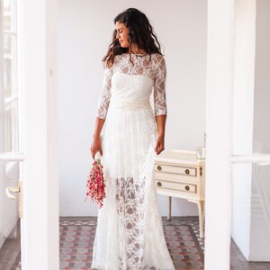 May include: A floor-length white lace dress featuring three-quarter length sleeves, a round neckline, and a solid white bodice underlay. The skirt has a sheer lace overlay with a scalloped hemline and a wide, textured waistband in a cream hue.