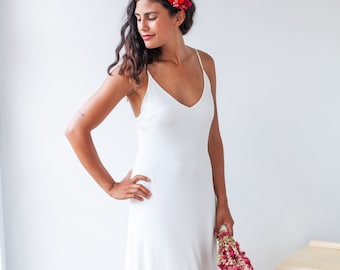 May include: A floor-length white slip dress with thin spaghetti straps and a V-neckline. The dress features a simple, fluid silhouette that falls straight to the floor. A small cluster of bright pink and red dried flowers is held in one hand.