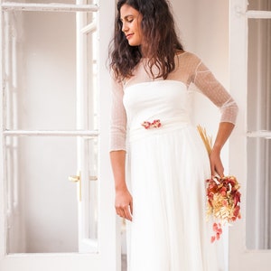May include: A floor-length white dress featuring a sheer overlay with a subtle dotted pattern, three-quarter length sleeves, and a wide waistband adorned with a cluster of dried red and orange flowers, paired with a matching dried floral bouquet.