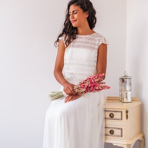 May include: A woman wearing a white lace crop top and a white tulle skirt. She is holding a bouquet of dried pink flowers and is sitting on a white stool in front of a white wall.