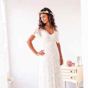 Woman in a bright room, next to a cream-y table decorated with flowers. She is dressed like a bride, wearing an ivory lace bridal gown.