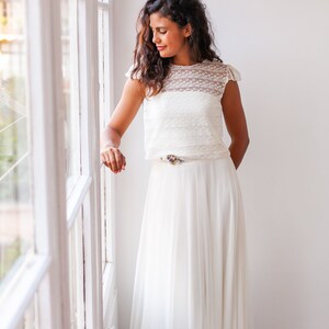 May include: A woman wearing a white lace top and a white tulle skirt. She is standing in front of a window and looking down.