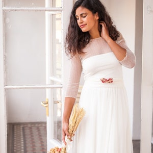 May include: A white floor-length dress featuring a sheer dotted mesh overlay on the bodice and three-quarter length sleeves, a solid white waistband, and a floral accent at the waist. The text www.mimetikbcn.com is printed at the bottom.