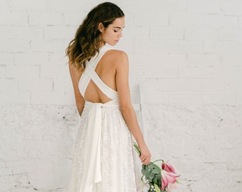 May include: A floor-length white dress featuring a lace skirt and a solid white bodice with wide straps that cross over the open back. The dress has a long fabric tie at the waist and a full-length skirt with a delicate floral lace overlay pattern.