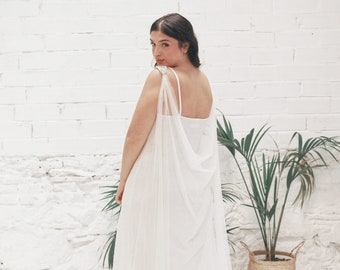 May include: A white wedding dress with a sheer cape overlay. The dress has a simple, elegant design and is perfect for a beach wedding.