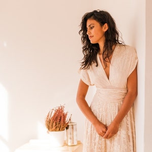 A woman wears a wedding dress with white walls and a chest of drawers at the back. She is wearing a boho-chic wedding dress. The dress is full made in a champagne satin jersey base and a champange lace overlay. The ambience is dreamy.