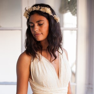 May include: An off-white sleeveless dress featuring a deep V-neckline, a wrapped bodice with horizontal fabric bands at the waist, and an overlay of delicate floral lace. A matching floral crown rests on the head. Text reads www.mimetikbcn.com.