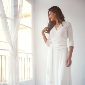 Woman wearing a simple white wrap dress as a wedding dress. The bridal gown is long and has a deep v-neck. She is staring outside while smiling.