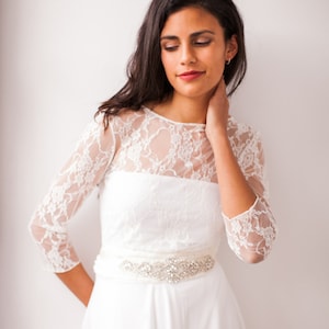 Könnte beinhalten: Ein bodenlanges weißes Kleid mit einem transparenten Spitzenoberteil, Dreiviertelärmeln und einem hohen Ausschnitt. Die Taille ist mit einem breiten, horizontalen Band akzentuiert, das mit funkelnden, klaren Strasssteinen und silberfarbenen Perlen besetzt ist.