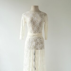 May include: A floor-length, off-white lace dress featuring long sleeves, a boat neckline, and a fitted waist sash. The garment is crafted from a sheer, intricate floral lace pattern that extends from the bodice down to the hemline.
