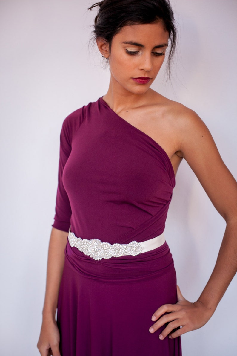 Puede incluir: Un vestido de un solo hombro en color ciruela intenso con un corpi&ntilde;o ajustado y tela fruncida en la cintura. Una faja ancha de sat&eacute;n blanco est&aacute; atada a la cintura, adornada con un panel decorativo central de pedrer&iacute;a y cuentas transparentes.