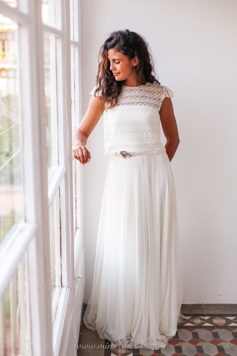 May include: A woman wearing a white lace top and a white tulle skirt. She is standing in front of a window and looking down.