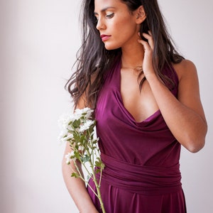 Puede incluir: Un vestido sin mangas de color ciruela intenso con un escote drapeado tipo capucha y una cintura ancha y ajustada que se envuelve alrededor del torso. El vestido tiene una falda fluida y se sostiene junto a un tallo de margaritas blancas.