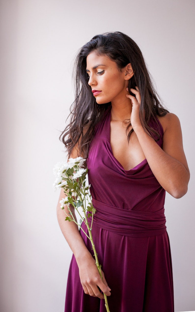 Puede incluir: Un vestido sin mangas de color ciruela intenso con un escote drapeado tipo capucha y una cintura ancha y ajustada que se envuelve alrededor del torso. El vestido tiene una falda fluida y se sostiene junto a un tallo de margaritas blancas.