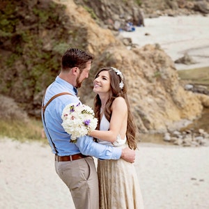 A couple of newly-married is on the beach. They look really happy and in love. The girl is wearing a big natural bouquet matching her flower crown in the hair. She wears a rustic bridal gown with ivory base and golden lace skirt.