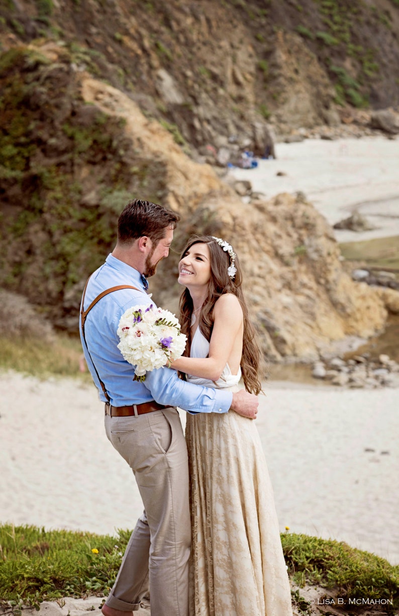 A couple of newly-married is on the beach. They look really happy and in love. The girl is wearing a big natural bouquet matching her flower crown in the hair. She wears a rustic bridal gown with ivory base and golden lace skirt.