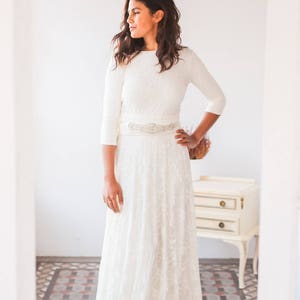 Woman is standing in a small room with natural light. She is wearing a long sleeve wedding dress. Her hand is on the waist and the other is relaxed over her overskirt