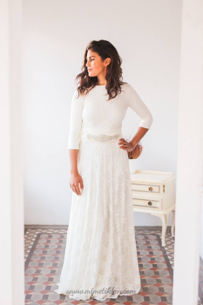 Woman is standing in a small room with natural light. She is wearing a long sleeve wedding dress. Her hand is on the waist and the other is relaxed over her overskirt