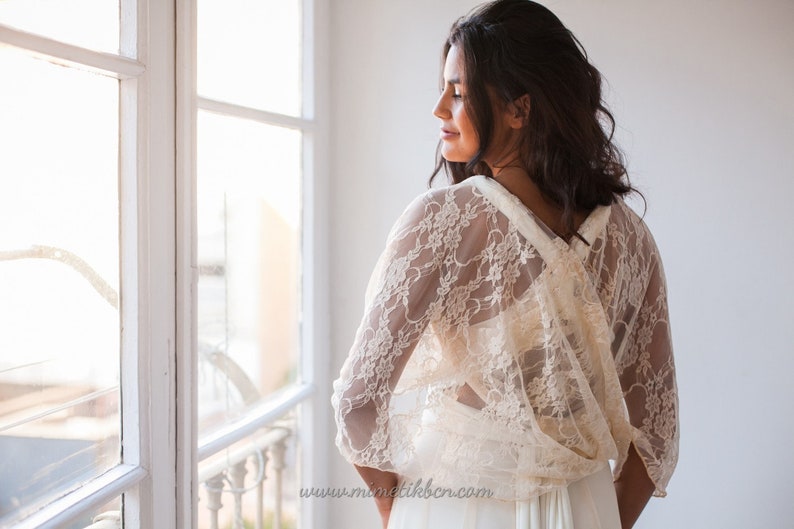 May include: An ivory lace bolero with three-quarter length sleeves and a delicate floral pattern. The garment features a draped back design with a crossover detail, worn over a plain white dress. The text www.mimetikbcn.com is visible.