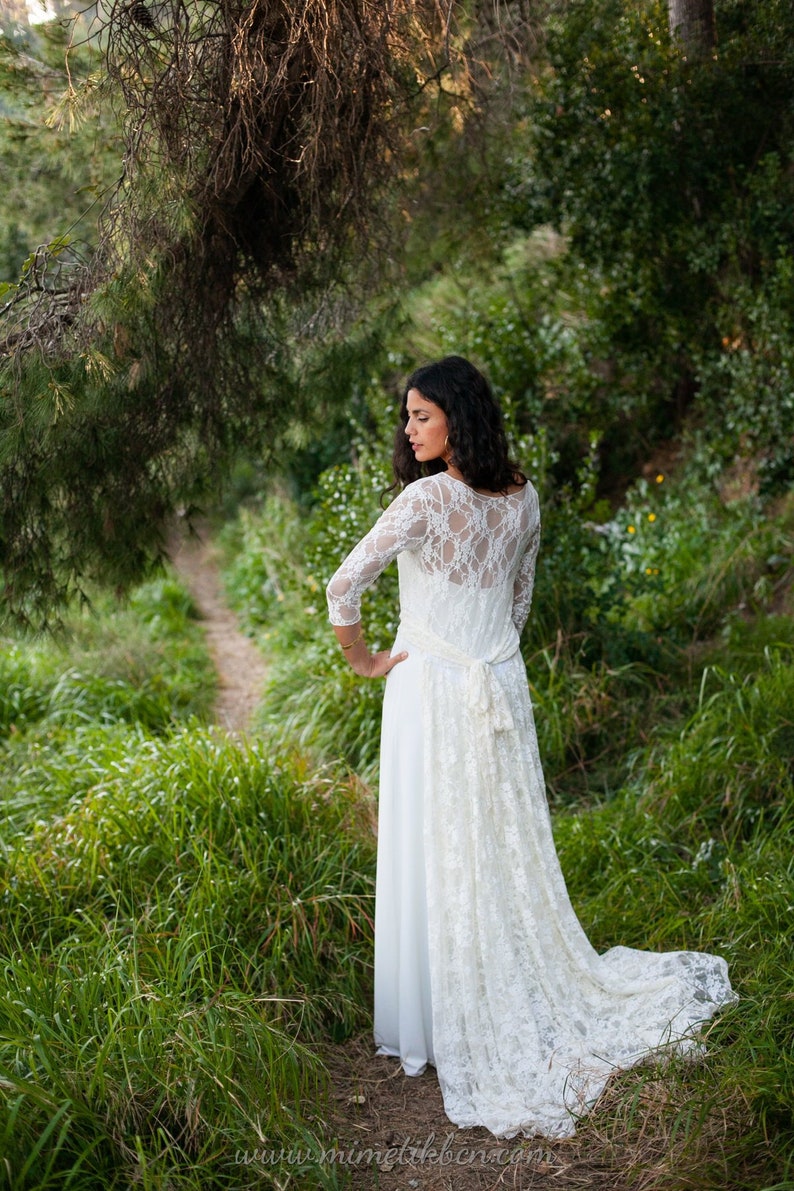May include: A floor-length white dress featuring three-quarter length lace sleeves, a sheer lace back panel, and a long lace train that attaches at the waist with a tie. The dress has a solid white skirt and a delicate floral lace overlay.
