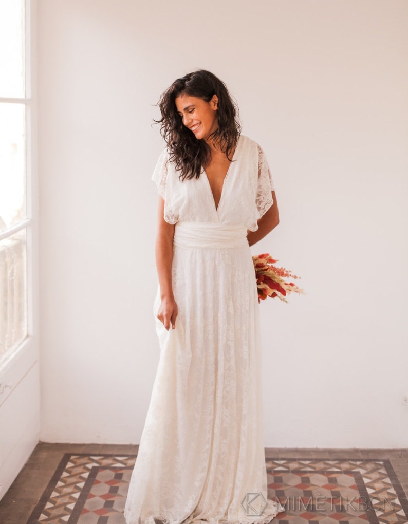 A woman wears a wedding dress with white walls at the back and hydraulic floor. She is wearing a femenine wedding dress made of ivory lace. She also wears a bouquet in the hands, which are in her back. She has long dark boho hair.