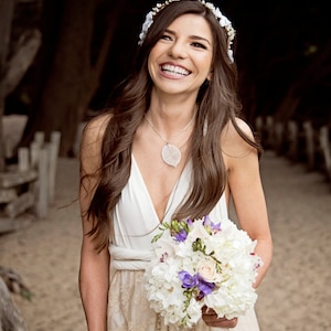 May include: A white sleeveless dress with a lace skirt, a silver leaf pendant necklace, and a floral headpiece. The outfit is paired with a bouquet of white hydrangeas and purple freesia flowers wrapped in a white ribbon with a rhinestone band.