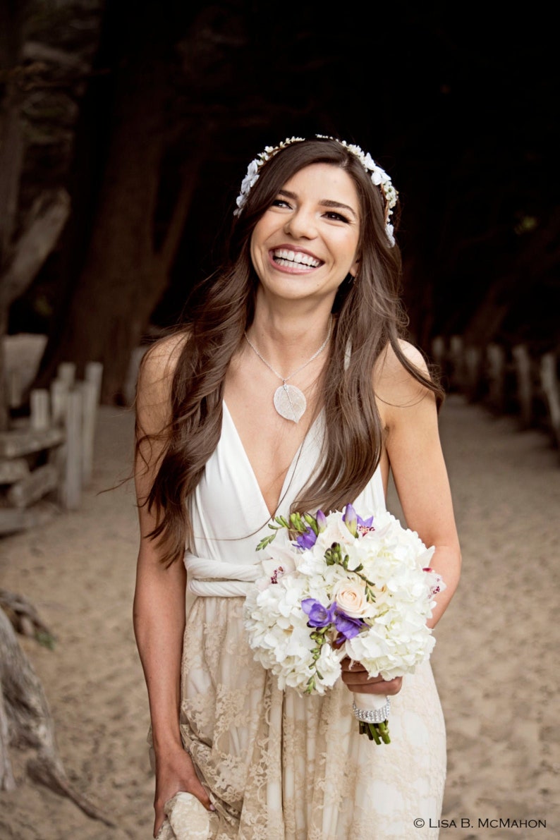 May include: A white sleeveless dress with a lace skirt, a silver leaf pendant necklace, and a floral headpiece. The outfit is paired with a bouquet of white hydrangeas and purple freesia flowers wrapped in a white ribbon with a rhinestone band.