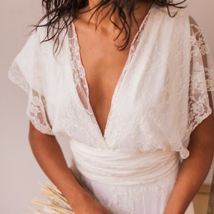 May include: A white lace dress featuring a deep V-neckline, short flutter sleeves, and a wide fabric waistband. The bodice and sleeves are crafted from floral lace, while the skirt is made of a matching white fabric. The text MIMETIKBCN is visible.