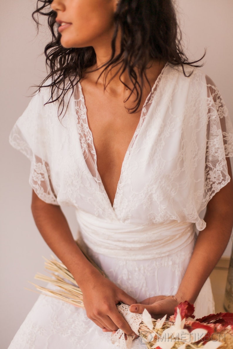 May include: A white lace dress featuring a deep V-neckline, short flutter sleeves, and a wide fabric waistband. The bodice and sleeves are crafted from floral lace, while the skirt is made of a matching white fabric. The text MIMETIKBCN is visible.