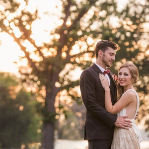 May include: A couple standing together outdoors at sunset. One individual wears a dark suit with a red bowtie, while the other wears a white sleeveless dress with a textured skirt. The text Feather Print Photography is visible in the bottom right corner.