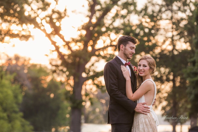 May include: A couple standing together outdoors at sunset. One individual wears a dark suit with a red bowtie, while the other wears a white sleeveless dress with a textured skirt. The text Feather Print Photography is visible in the bottom right corner.