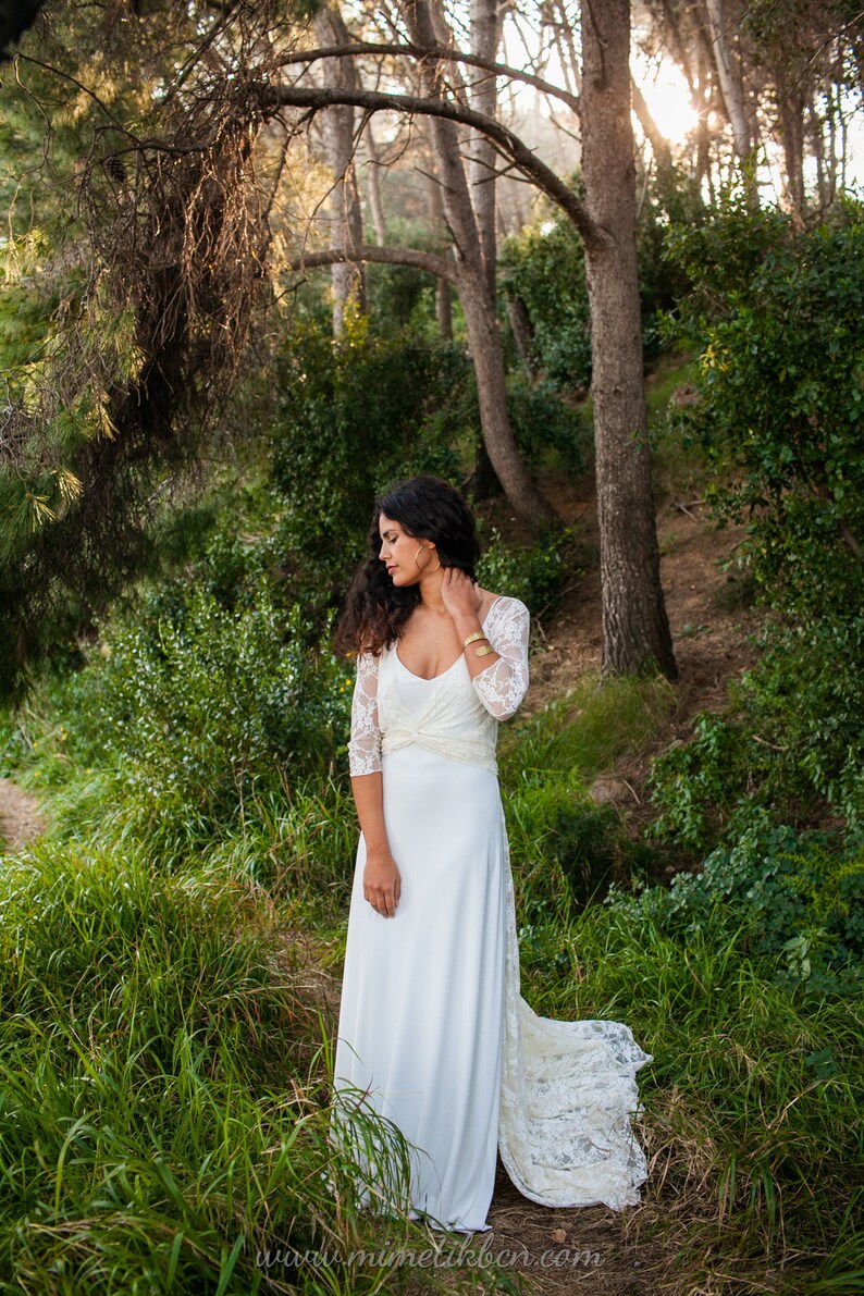 May include: A floor-length white dress featuring three-quarter length lace sleeves, a scoop neckline, and a gathered waist detail. The bodice is constructed from lace, while the skirt is made of a smooth, solid white fabric with a lace train.
