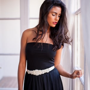 May include: A woman wearing a black strapless dress with a white rhinestone belt. She is standing by a window and looking down.
