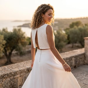 Könnte beinhalten: Ein weißes zweiteiliges Kleid bestehend aus einem ärmellosen bauchfreien Oberteil mit texturiertem Paillettenbesatz und offenem Rücken. Der passende bodenlange Rock hat eine hohe Taille und eine vertikale Reihe kleiner runder Knöpfe am Rücken.