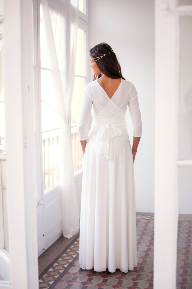 The woman is showing her back to the viewer. She is wearing an ivory satin jersey wedding dress with three-quarter sleeves. She wears vintage jewelry on her hair.